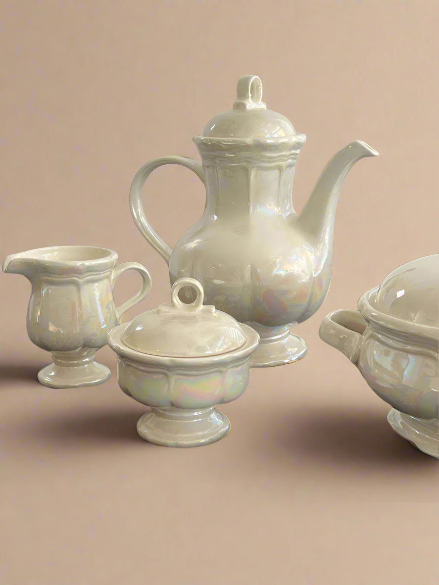 Set of white ceramic teapot, creamer, sugar bowl, and teacup on a black surface with a white background.