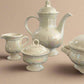 Set of white ceramic teapot, creamer, sugar bowl, and teacup on a black surface with a white background.